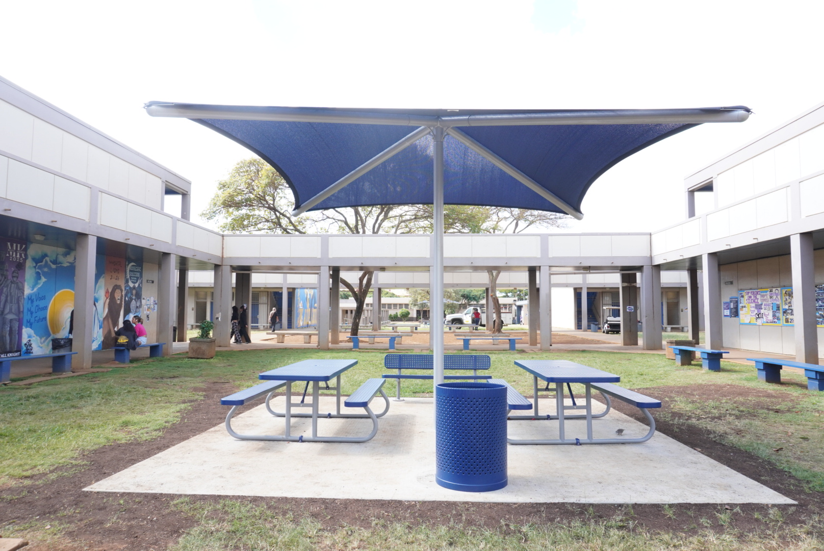 Pictured is one out of five different sets of umbrella covered tables. This set can be found in between G and H buildings! It is located off the pathway to the counselors breezeway and can be easily seen from the class murals. 